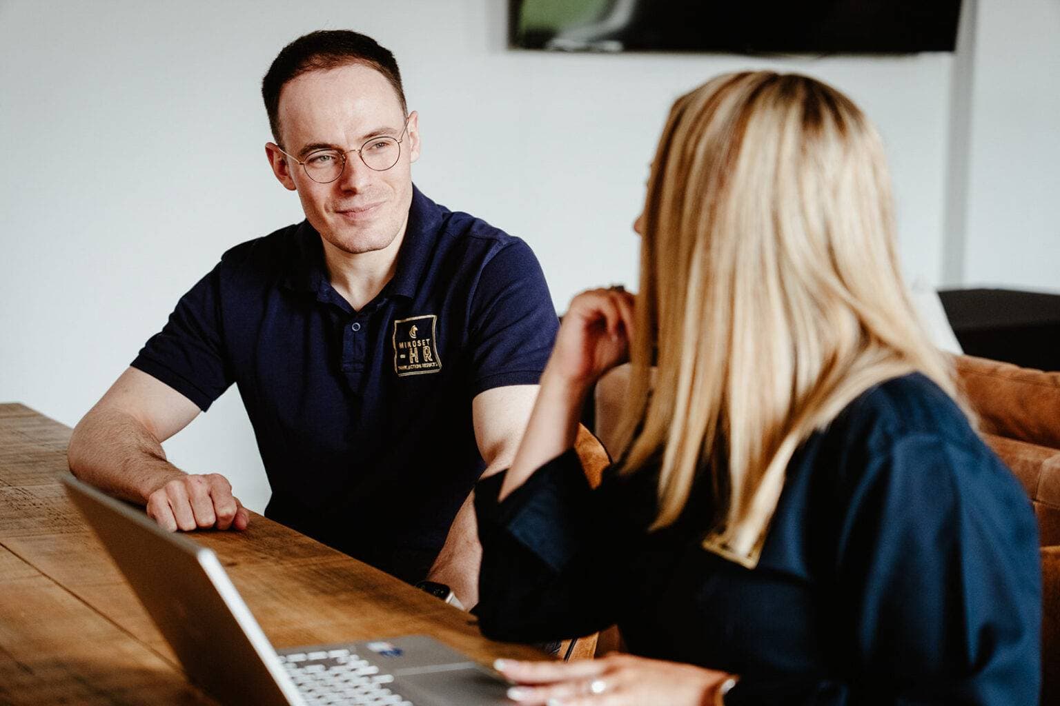 A Mindset HR consultant in a branded polo shirt in conversation with a client across a wooden table, a laptop open between them